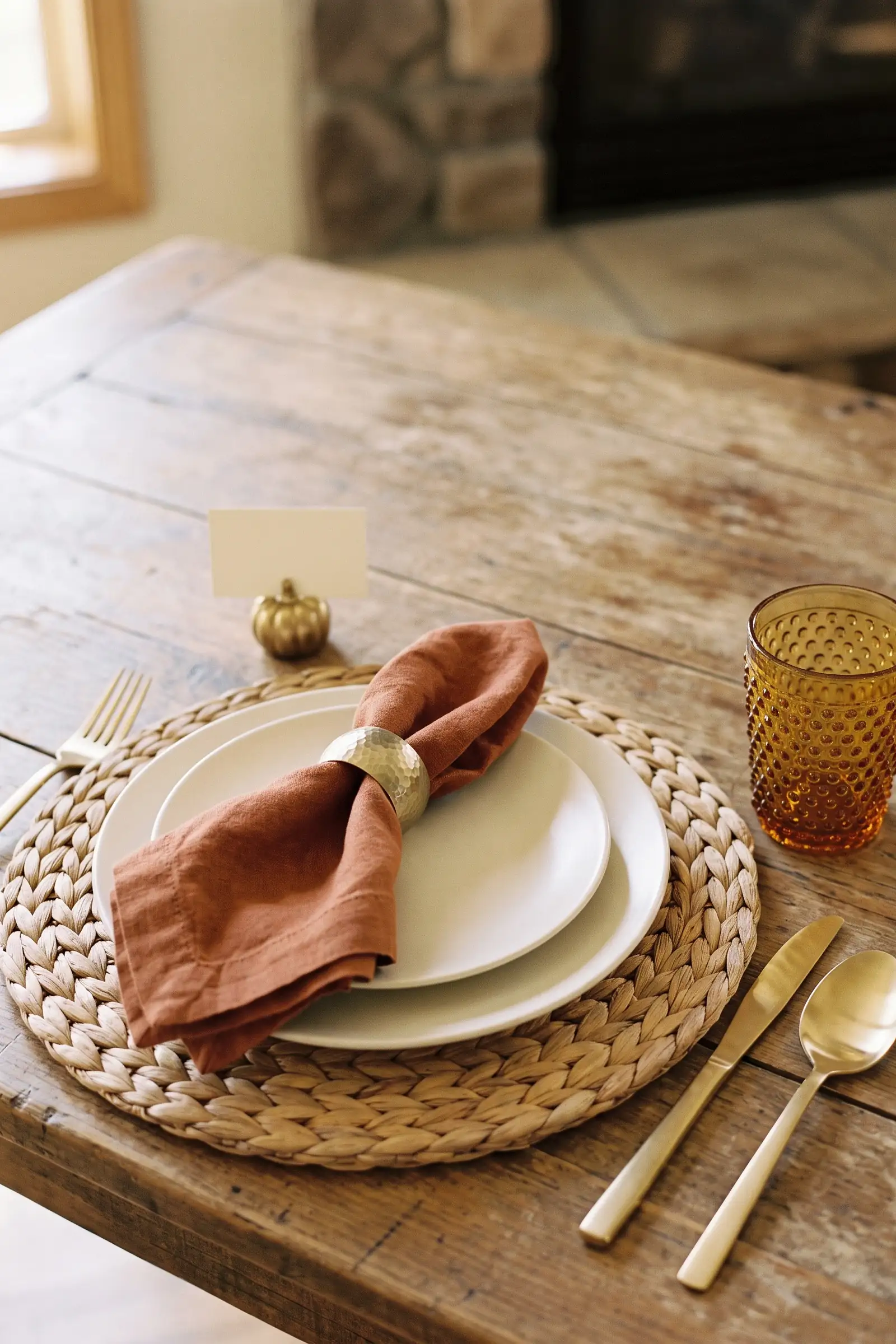Thanksgiving place setting with woven placemat, ivory plates, rust napkin, gold ring, and amber glass